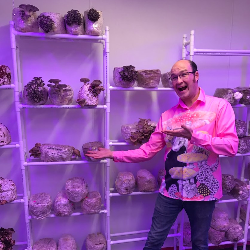 A shelf of PVC pipes stands before a white wall in purple light. Avery Stempel stands before it, gesturing like Vanna White, marveling at the fruiting mushrooms on the racks of the shelves. He is wearing a button down shirt with mushrooms and cats.