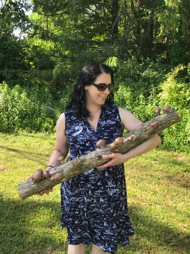 A field with a forest beyond it. In the foreground is Amy Hood, cradling a large log with fungi emerging from it.